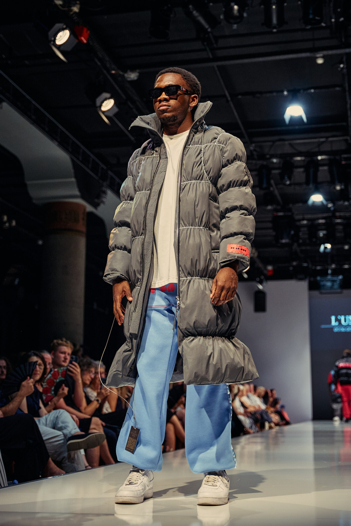 Look 02 „La Réflexion“ — langer grauer Reflex-Puffermantel über weißem Shirt, hellblaue weite Hose und weiße Sneaker auf der Mannheimer Runway
