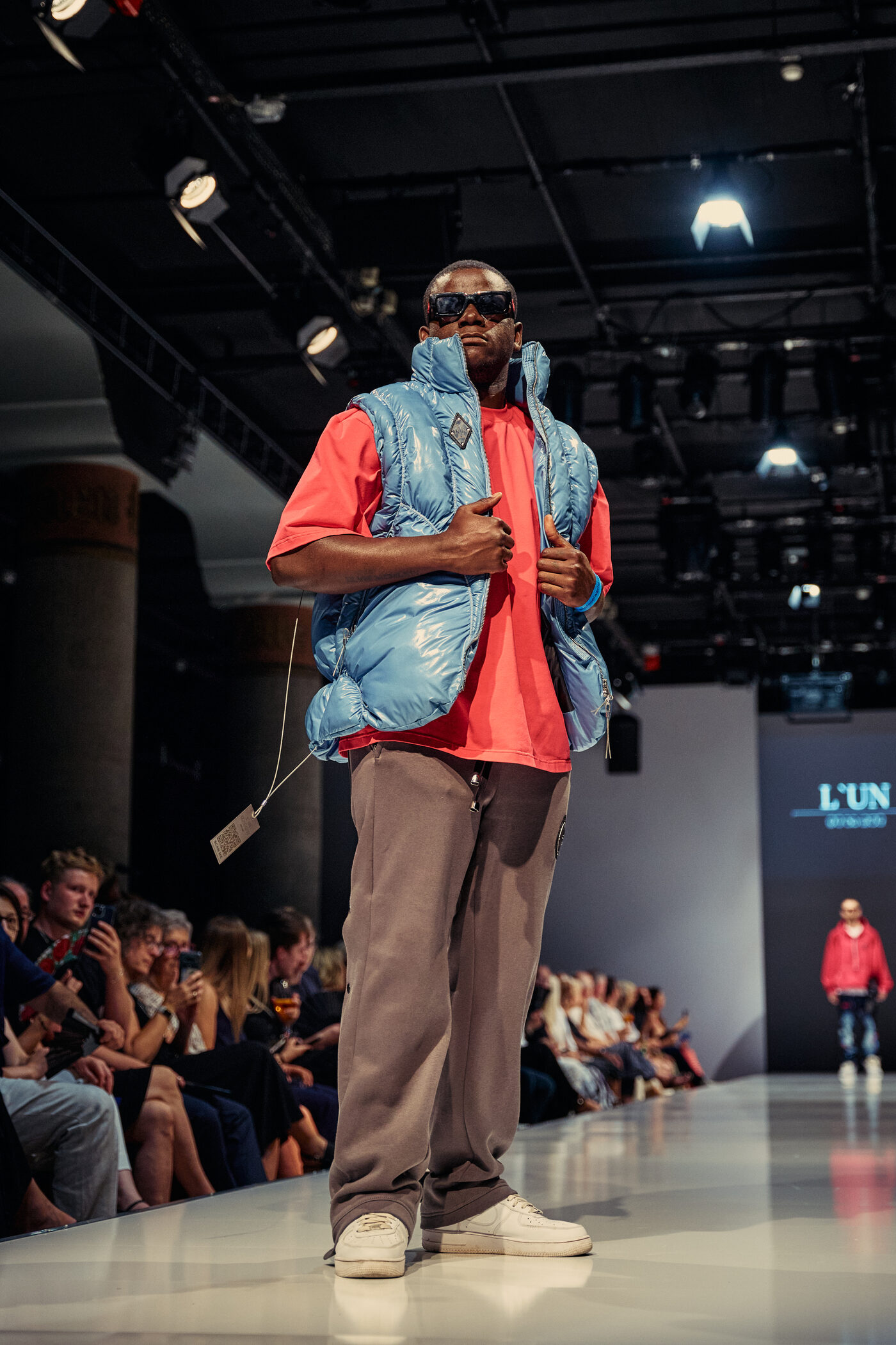 Look 03 „Éclat Blue“ — hellblaue Glanz-Pufferweste über korallrotem Oversize-Shirt, taupefarbene weite Hose und weiße Sneaker auf der Mannheimer Runway