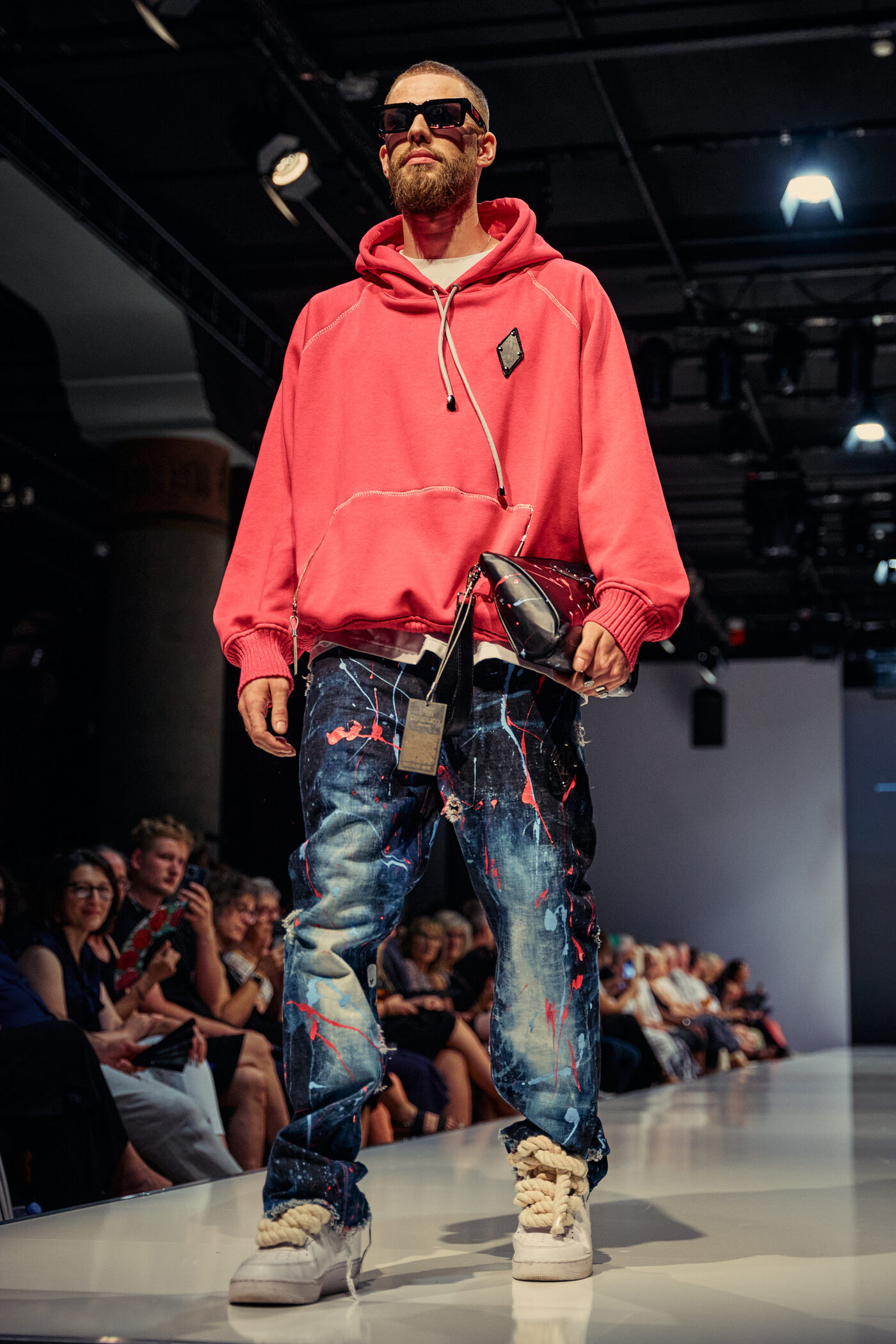 Look 04 „Le Calme“ — korallroter Oversize-Hoodie, farbbespritzte Baggy-Jeans, weiße Sneaker mit Kordel-Schnürung und glänzende Clutch auf der Mannheimer Runway
