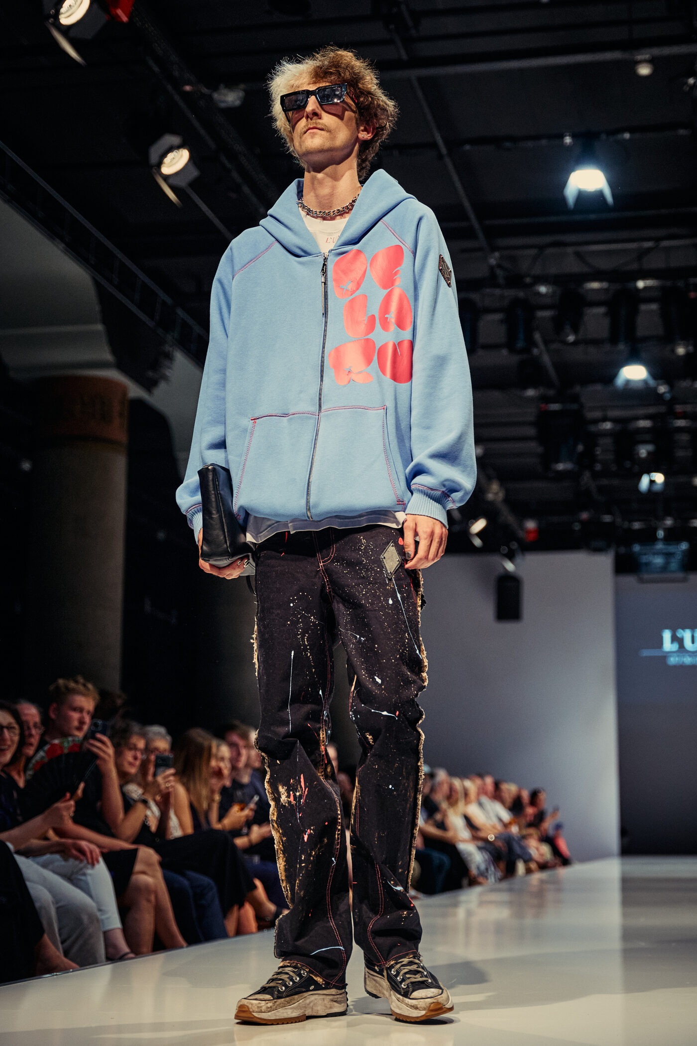 Look 05 „Le Fort“ — hellblauer Zip-Hoodie mit rotem Graffiti-Print, schwarze farbbespritzte Used-Jeans, getragene Sneaker und schwarze Clutch auf der Mannheimer Runway
